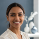 Headshot of a smiling South Asian woman, mid-20s to early 30s, against a blurred slate blue and white background.